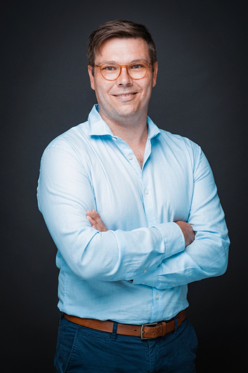 Man wearing a light blue button-up shirt and glasses smiling with arms crossed